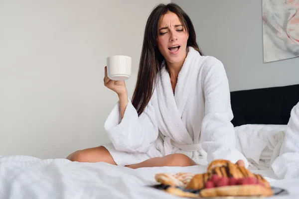 젊은 매력적인 여자 와 어두운 머리 에 흰 목욕 가운 들고 컵 커피 손에 놀랍게도 찾고 맛있는 과자 에 있는 아침 식사 에 침대에서 현대 호텔 — 스톡 사진