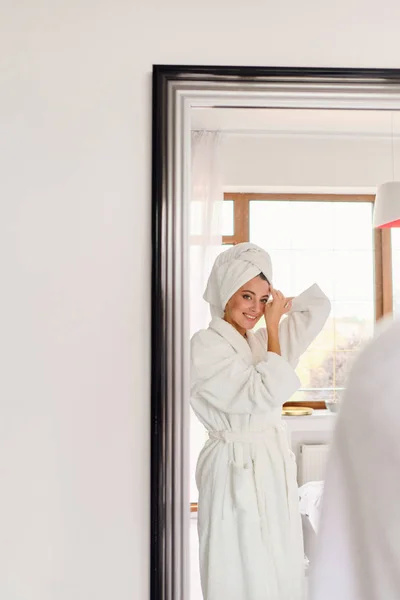 Jeune belle femme souriante en peignoir blanc et serviette sur la tête regardant joyeusement dans le miroir à l'hôtel confortable moderne — Photo