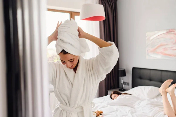 Joven mujer sonriente atractiva en albornoz blanco y toalla en la cabeza mirando soñadoramente hacia abajo, mientras que la chica bonita acostada en la cama sobre el fondo en el moderno y acogedor hotel —  Fotos de Stock