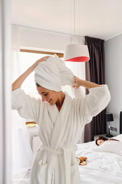 Jeune jolie femme souriante en peignoir blanc et serviette sur la tête regardant joyeusement vers le bas tandis que la fille amie couchée dans le lit sur le fond dans un hôtel confortable moderne — Photo
