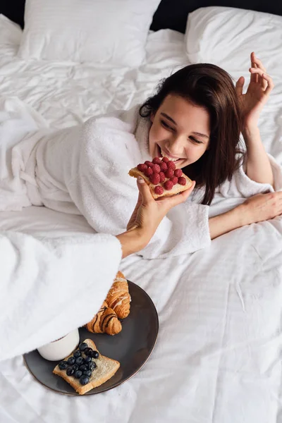 Jeune femme joyeuse en peignoir blanc couché au lit et manger avec un ami délicieuse pâtisserie sur le petit déjeuner à l'hôtel moderne confortable — Photo