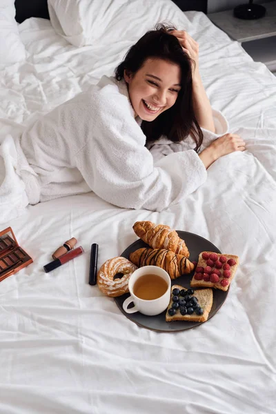 Fiatal, gyönyörű nevető nő, fehér fürdőköpenyben boldogan az ágyban fekve, finom péksüteményekkel és egy csésze tea reggelivel a modern, otthonos szállodában — Stock Fotó