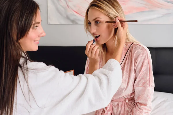 Young attractive woman with blond hair in pink velvet robe dreamily looking at pretty girl that happily doing makeup to her on big bed in modern cozy hotel — Stock Photo, Image