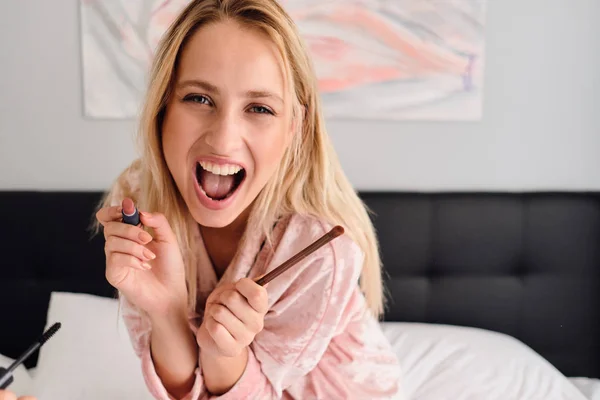 Joven mujer alegre con el pelo rubio en bata de terciopelo rosa sosteniendo lápiz labial y pequeño cepillo en las manos mirando alegremente en la cámara sentada en la cama en el hotel moderno — Foto de Stock