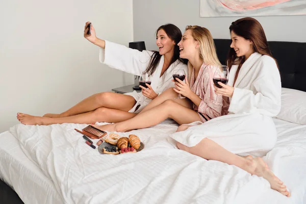 Grupo de jovens mulheres sorridentes em roupões de banho segurando copos com vinho tinto nas mãos felizes tirando fotos no celular juntos. Meninas bonitas tomando café da manhã na cama no hotel moderno — Fotografia de Stock