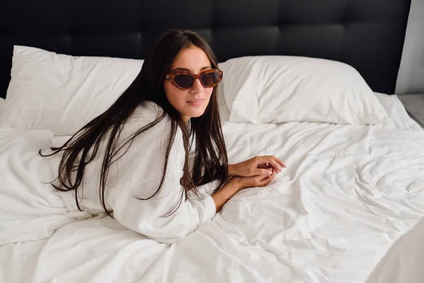 Молодая красивая женщина с темными волосами в белом халате и солнцезащитных очках мечтательно смотрит в камеру лежащей на большой кровати в современном отеле — стоковое фото