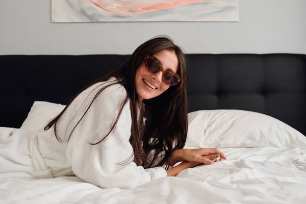 Fiatal szép mosolygó nő Sötét haja fehér fürdőköpeny és napszemüveg örömmel keres kamera fekszik a nagy ágy, modern hangulatú szálloda — Stock Fotó