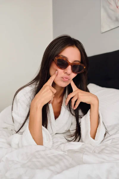 Mujer atractiva joven con cabello oscuro en albornoz blanco y gafas de sol que sostiene cuidadosamente los dedos cerca de las mejillas tumbadas en la cama grande en el moderno y acogedor hotel — Foto de Stock