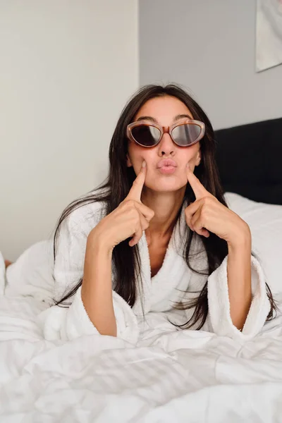 Young beautiful woman with dark hair in white bathrobe and sunglasses holding fingers near cheeks dreamily looking in camera lying on big bed in modern cozy hotel — Stock Photo, Image
