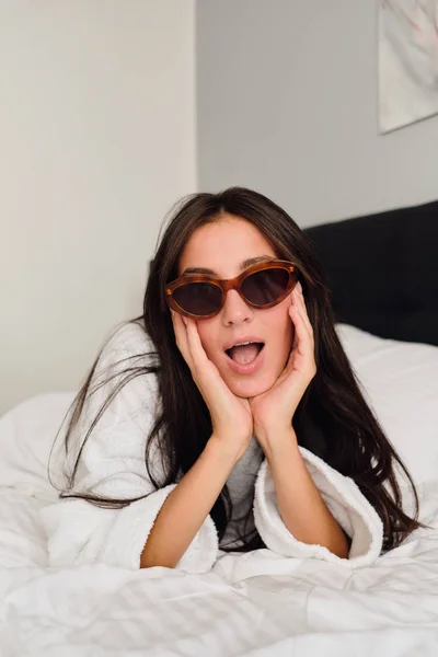 Young attractive woman with dark hair in white bathrobe and sunglasses leaning on hands dreamily looking in camera on big bed in modern hotel — Stock Photo, Image