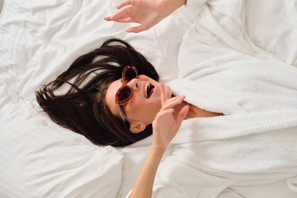Joven mujer sonriente emocional con cabello oscuro en albornoz blanco y gafas de sol mirando alegremente a un lado tumbado en la cama en el moderno y acogedor hotel — Foto de Stock