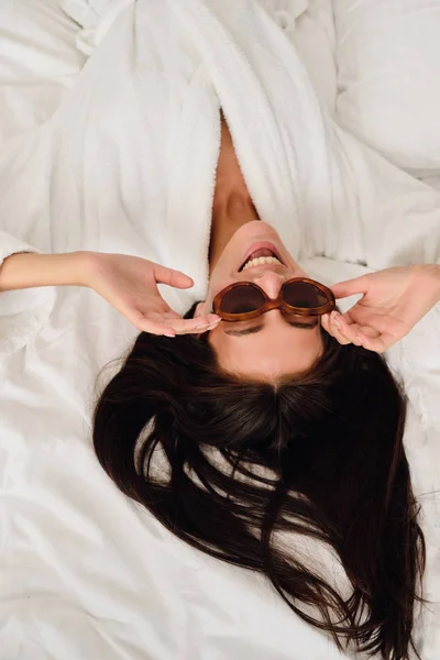 Young beautiful smiling woman with dark hair in white bathrobe and sunglasses happily lying on big bed in modern cozy hotel — Stock Photo, Image