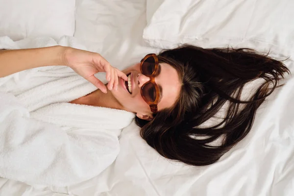 Joven hermosa mujer sonriente con cabello oscuro en albornoz blanco y gafas de sol dedo mordedura de ensueño acostado en la cama en el moderno y acogedor hotel — Foto de Stock