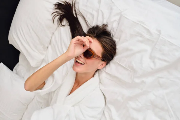 Joven hermosa mujer sonriente con cabello oscuro en albornoz blanco y gafas de sol sosteniendo la mano cerca de la cara felizmente tumbado en la cama grande en el hotel moderno — Foto de Stock
