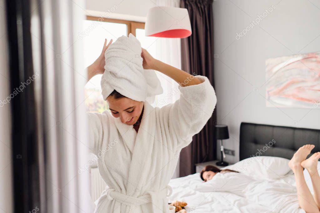 Young attractive smiling woman in white bathrobe and towel on head dreamily looking down while pretty girl lying in bed on background in modern cozy hotel