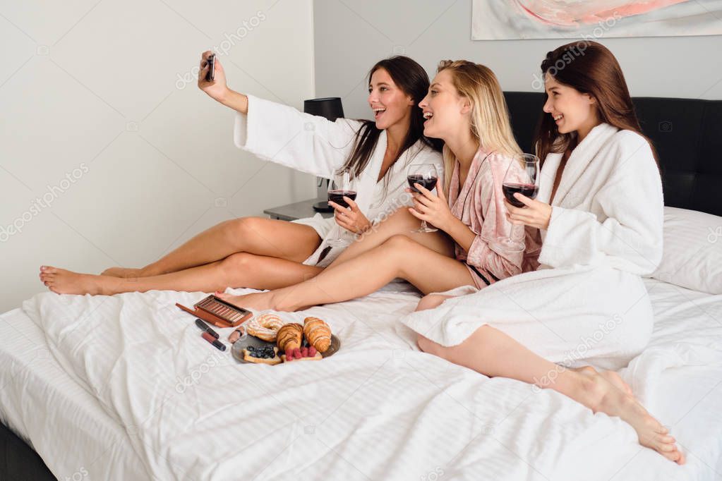 Group of young smiling women in bathrobes holding glasses with red wine in hands happily taking photos on cellphone together. Beautiful girls having breakfast in bed in modern hotel