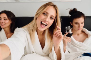 Beyaz bornozlu sarı saçlı genç neşeli kadın elinde göz fırçası ile kameraya mutlu bakarken, arka plandaki kızlar modern otelde yatakta şarapla hayali olarak oturuyorlar.