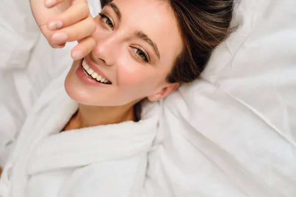 Young gorgeous smiling woman with dark hair in white bathrobe dreamily looking in camera lying on bed in cozy hotel — Stock Photo, Image