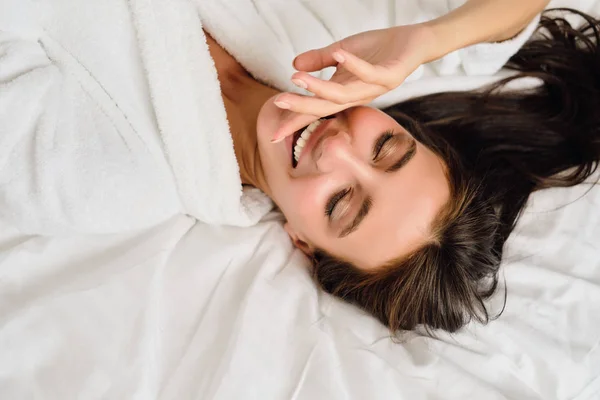 Joven y atractiva mujer sonriente con cabello oscuro en albornoz blanco cogida de la mano cerca de la boca acostada en la cama en un moderno y acogedor hotel — Foto de Stock