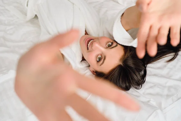 Junge schöne Frau mit dunklen Haaren im weißen Bademantel hält die Hände hoch und schaut glücklich in die Kamera, wie sie im gemütlichen Hotel auf dem Bett liegt — Stockfoto