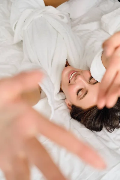 Joven y atractiva mujer sonriente con cabello oscuro en albornoz blanco cubriendo la cámara con la mano cerrando los ojos soñadoramente tumbada en la cama en el acogedor hotel — Foto de Stock