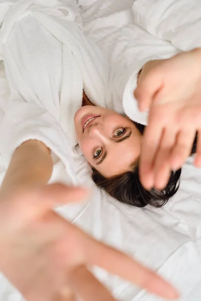 Young attractive woman with dark hair in white bathrobe holding hands up dreamily looking in camera lying on bed in cozy hotel — Stock Photo, Image