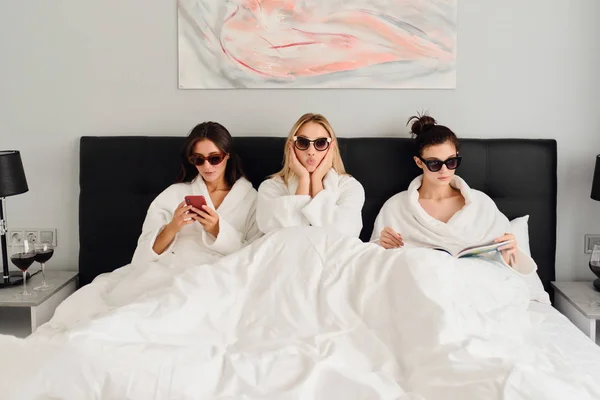 Три молодих красивих жінки в білих халатах і сонцезахисних окулярах лежать в ліжку в сучасному затишному готелі. Красива дівчина мрійливо дивиться в камеру, в той час як дві дівчини поруч використовують мобільний телефон і читають книгу — стокове фото