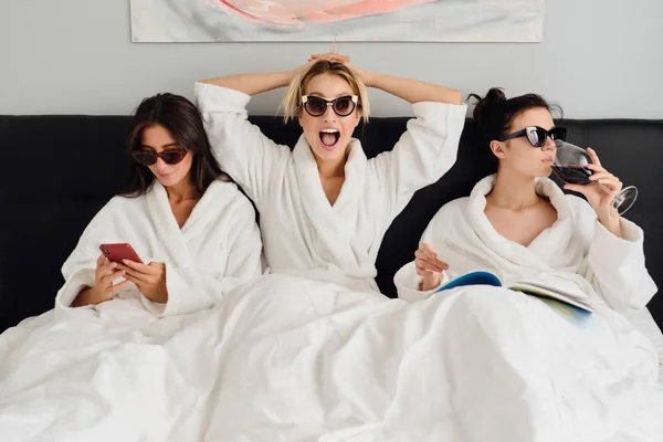 白いバスローブとサングラスを着た3人の若い魅力的な女性が、モダンな居心地の良いホテルのベッドに横たわっています。携帯電話を使い、ワインで本を読む2人の女の子が近くにいる間、女の子は感情的にカメラを見ている — ストック写真