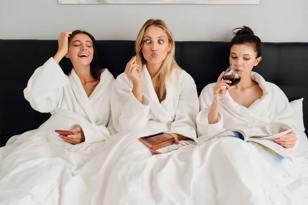 モダンな居心地の良いホテルでベッドに横たわっている白いバスローブを着た3人のかわいい女性。かわいい女の子は、携帯電話を使用して楽しく近い女の子がワインで本を読みながら、手にアイシャドウパレットを保持している顔を作ります — ストック写真