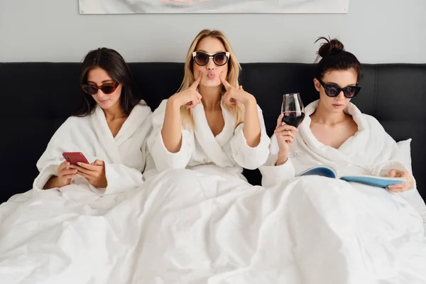 Három fiatal, gyönyörű nő fehér fürdőköpenyben és napszemüveg az ágyban fekve, modern hangulatú szállodában. Csinos lány játékosan keresett a kamera, míg a két lány közelében használó cellphone és olvasási könyvet a bor — Stock Fotó