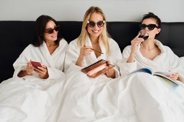 Três jovens mulheres bonitas em roupões de banho brancos e óculos de sol deitados na cama no moderno hotel acolhedor. Menina bonita segurando paleta sombra olho enquanto duas meninas perto de usar celular e livro de leitura com vinho — Fotografia de Stock