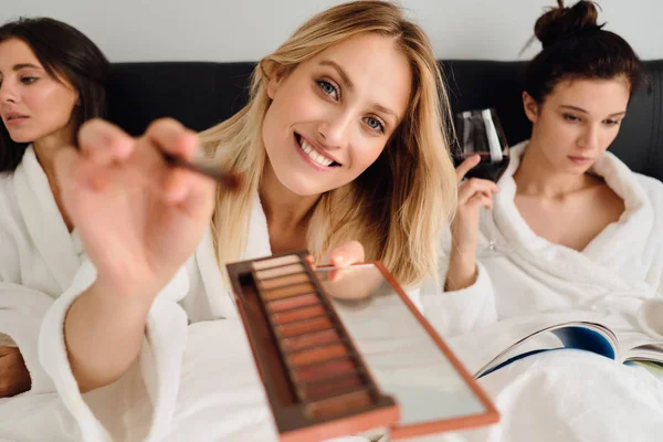 Jonge mooie lachende vrouw in witte badjas gelukkig op zoek in de camera met oogschaduw palet in de handen terwijl meisjes op de achtergrond dromerige zitten met wijn in bed in modern hotel — Stockfoto