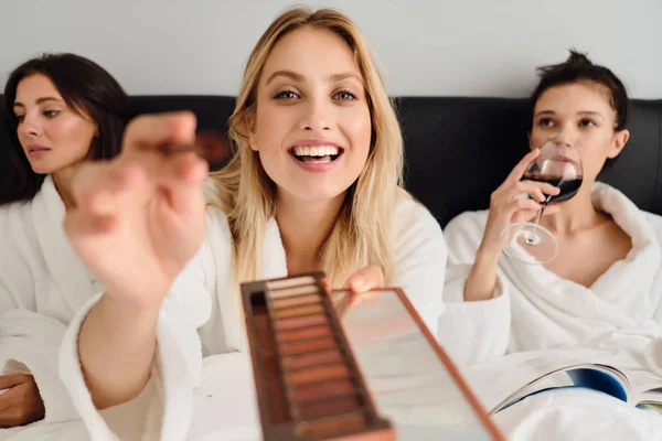 Jovem mulher sorridente bonita em roupão de banho branco feliz olhando na câmera com paleta de sombra nos olhos nas mãos, enquanto as meninas no fundo sentado com vinho na cama no moderno hotel acolhedor — Fotografia de Stock