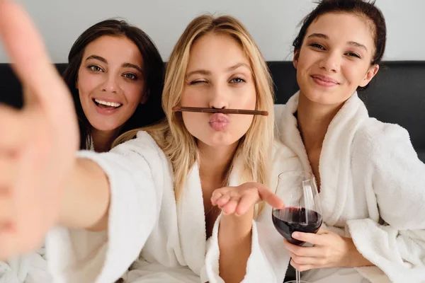 Mladá krásná žena se zasněně dívala do kamery s očima nad rty. Tři nádherné dívky v bílých županech s vínem v posteli šťastně fotí fotky v moderním hotelu — Stock fotografie