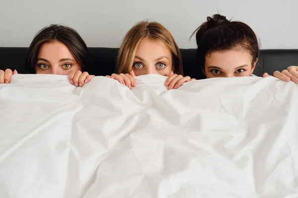 Три молодих красивих жінки сидять у ліжку і закривають обличчя білою ковдрою, мрійливо дивлячись в камеру разом в сучасному готелі — стокове фото