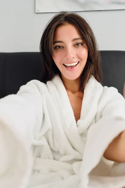 Jonge prachtige lachende vrouw in witte badjas zittend in bed gelukkig op zoek in de camera in het moderne hotel — Stockfoto