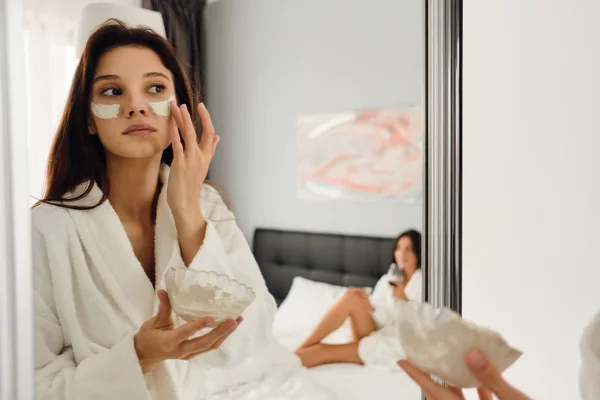 Jonge mooie vrouw in witte badjas maken cosmetische masker zorgvuldig op zoek in spiegel met meisje op achtergrond in modern hotel — Stockfoto