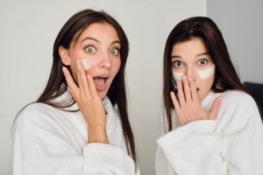 Beyaz bornozkoyu saçlı iki genç çekici kadın modern otelde kameraya bakarak yüzleri üzerinde kozmetik maskesi ile yatakta oturan 