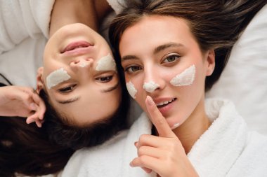 Beyaz bornozlu koyu saçlı iki genç çekici kadın, modern otelde dudakların yanında parmak tutan kameraya bakarak yüzleri üzerinde kozmetik maskesi ile yatakta yatan 