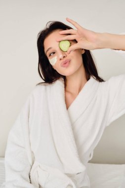 Beyaz bornozlu genç, yüzünde kozmetik maskesi olan genç, modern otelde hayali bir kenara bakan salatalıkla göz örtülüyorum 