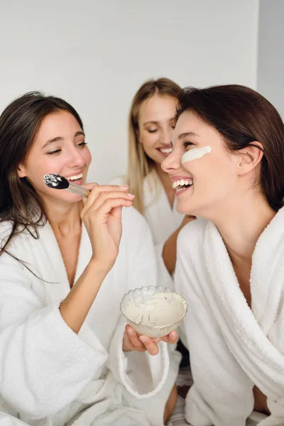 Tres mujeres jóvenes y alegres en batas blancas sentadas en la cama haciendo felizmente la máscara cosmética juntas en el hotel moderno —  Fotos de Stock