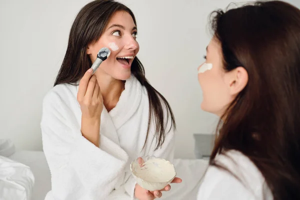 Joven hermosa mujer sonriente en albornoz blanco haciendo máscara cosmética felizmente mirando a un lado con chica bonita cerca en hotel moderno —  Fotos de Stock