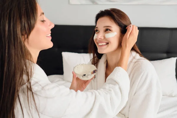 Jonge mooie lachende vrouw met donker haar in witte badjas zittend op bed gelukkig kijken opzij terwijl mooi meisje in de buurt van het maken van cosmetische masker haar in modern hotel — Stockfoto