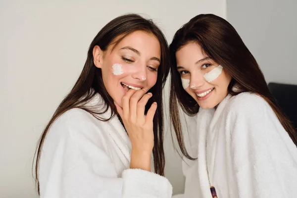 Dos mujeres hermosas jóvenes con el pelo oscuro en albornoces blancos sentados en la cama con máscara cosmética en caras que miran alegremente en cámara en hotel moderno —  Fotos de Stock
