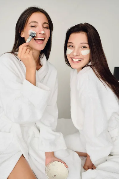 Two young beautiful smiling women with dark hair in white bathrobes sitting on bed with cosmetic mask on faces joyfully looking in camera in modern cozy hotel — Stock Photo, Image