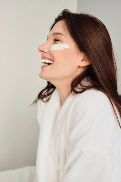 Yüzünde kozmetik maskesi ile beyaz bornoz genç neşeli kadın mutlu modern otelde kenara bakıyor — Stok fotoğraf