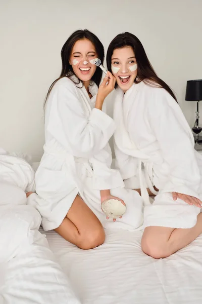 ベッドの上に座っている白いバスローブに黒髪の2人の若い美しい陽気な女性は、幸せにモダンなホテルで一緒に顔に化粧品のマスクを作ります — ストック写真