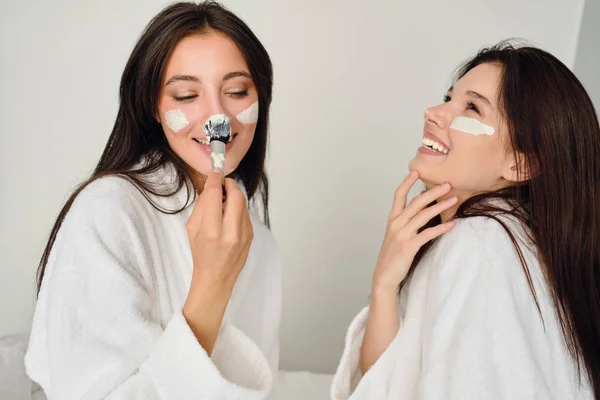 Dwie młode ładne uśmiechnięte kobiety z ciemnymi włosami w białych szlafrokach siedzących na łóżku z radością czyniąc maskę kosmetyczną na twarzach razem w nowoczesnym, przytulnym hotelu — Zdjęcie stockowe