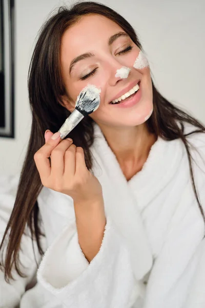 Joven mujer bastante sonriente en albornoz blanco haciendo máscara cosmética en la cara con cepillo felizmente cerrando los ojos en el hotel moderno —  Fotos de Stock