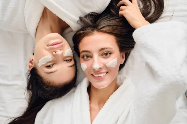 Twee jonge prachtige glimlachende vrouwen met donker haar in witte badjassen liggen in de buurt in bed met cosmetische masker op gezichten dromerige op zoek in de camera samen in moderne gezellige Hotel — Stockfoto